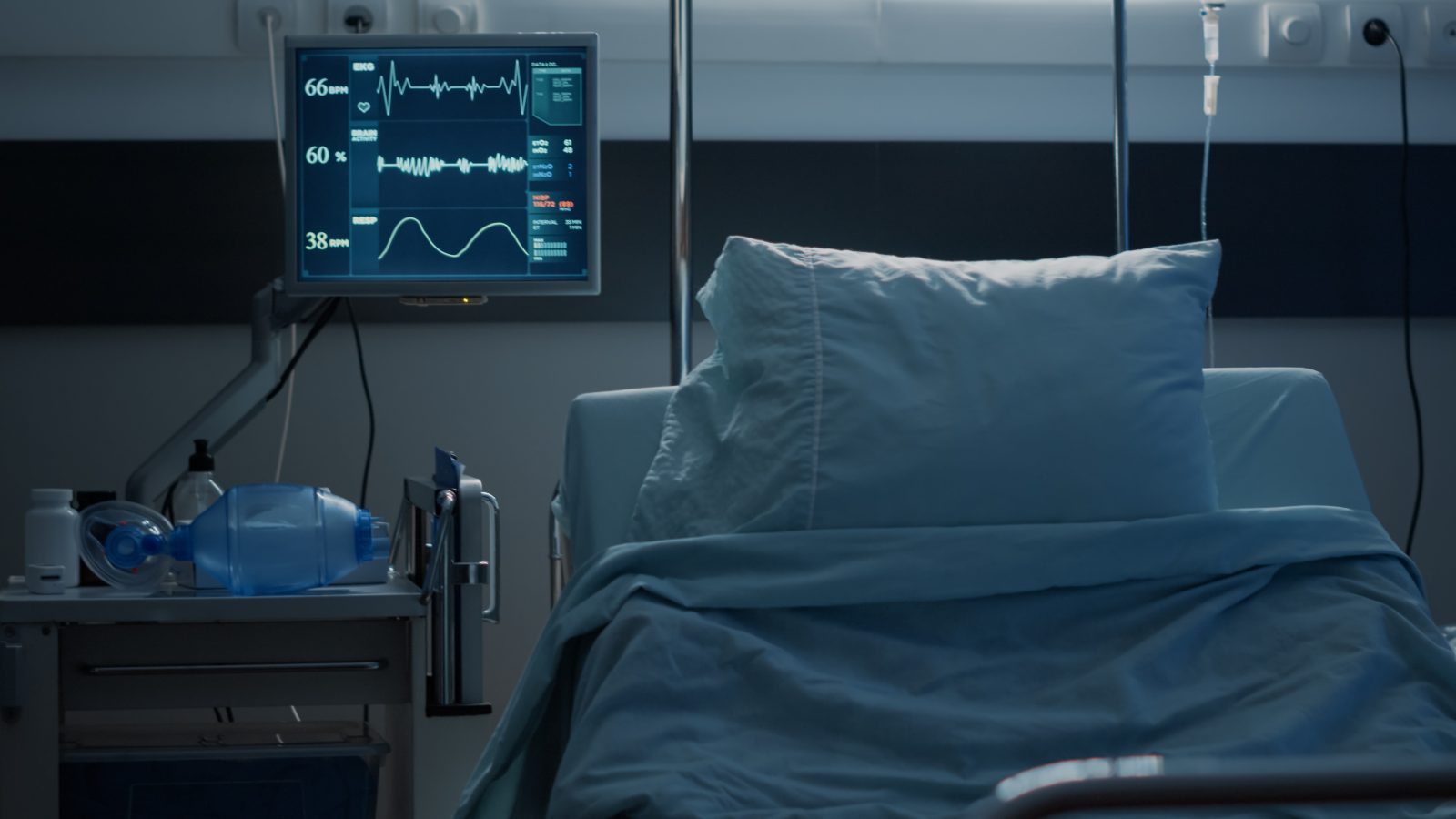 A dimly lit hospital room with an empty bed and a bedside monitor showing a heart rhythm trace.