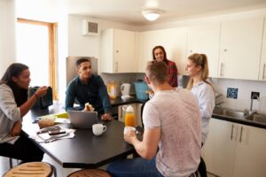 Several housemates standing and sitting around a kitchen island in a shared home, talking over coffee and juice.