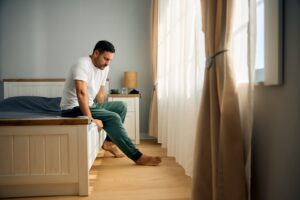 A man sitting on the edge of a bed in a bedroom, looking down, with morning light coming through the curtains.