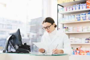A pharmacist in a white coat reviewing a prescription at a pharmacy counter, with shelves of medicine boxes behind her.