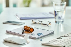 An open amber prescription bottle on a spiral notepad, with a few blue tablets, reading glasses and a glass of water on a bright desk.