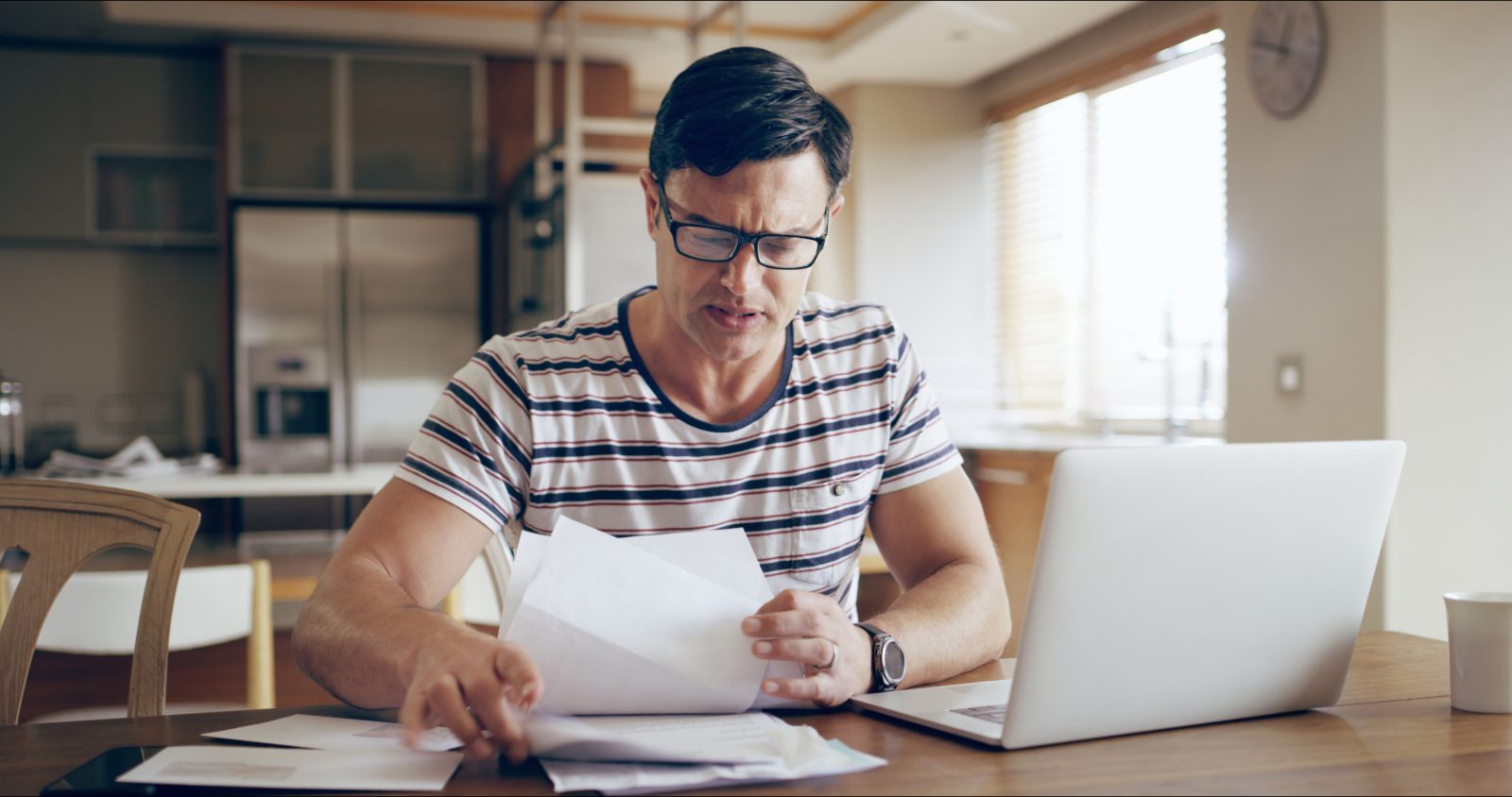 A man at a kitchen table reading a document, with a laptop and more paperwork spread in front of him.