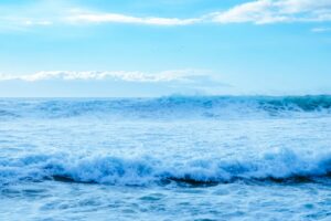 A wide view of the sea from the shoreline, with a line of waves breaking under an open sky.