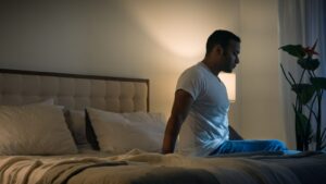 A man in a white T-shirt sitting on the edge of his bed at night, lit by a single soft lamp.
