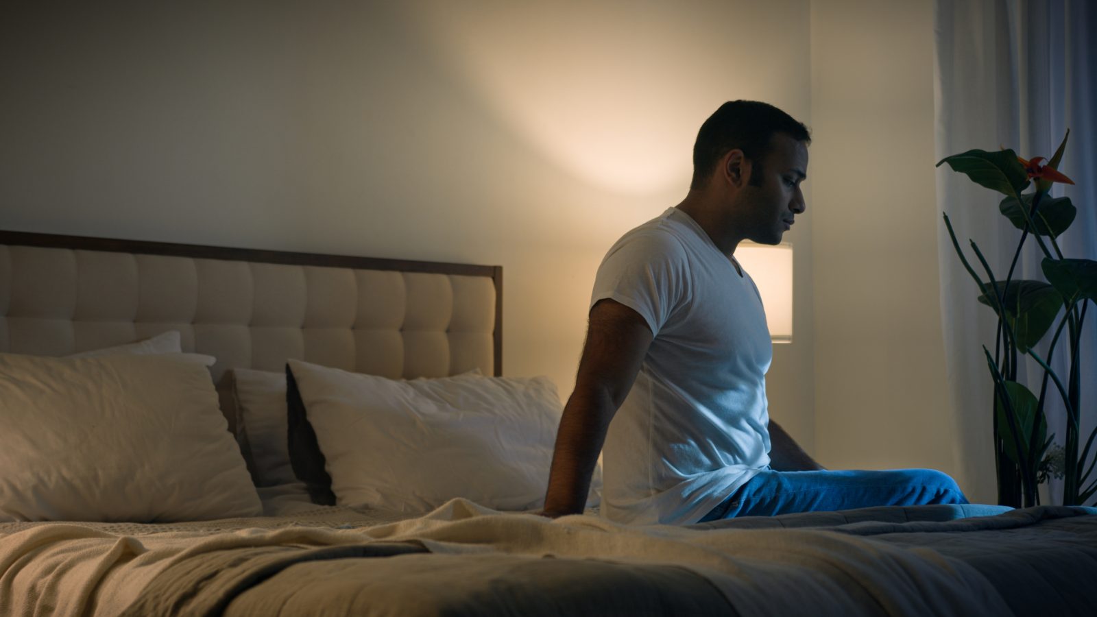 A man in a white T-shirt sitting on the edge of his bed at night, lit by a single soft lamp.