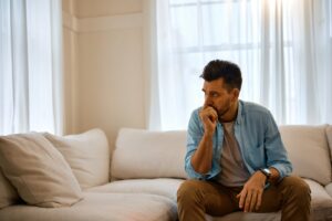 A man sitting on a light sofa in a bright living room, hand at his mouth, looking away in thought.
